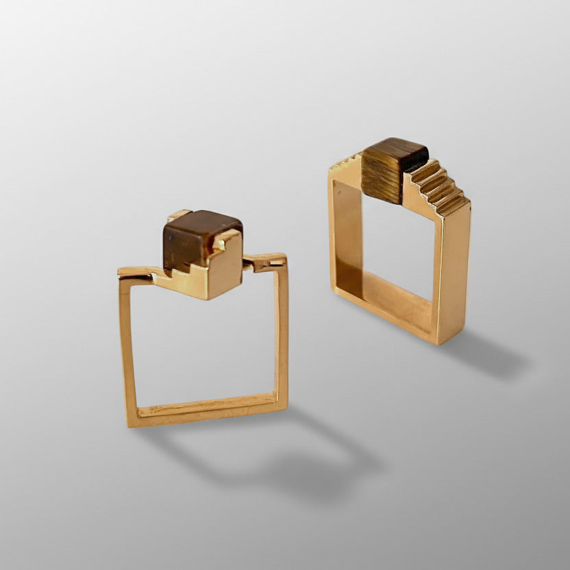 Deux bagues géométriques en vermeil, ornées d'un cube en oeil de tigre entre deux éléments en forme d'escalier. L'une des bagues est composée d'un anneau fin carré, tandis que l'autre est réalisée avec un anneau carré large.