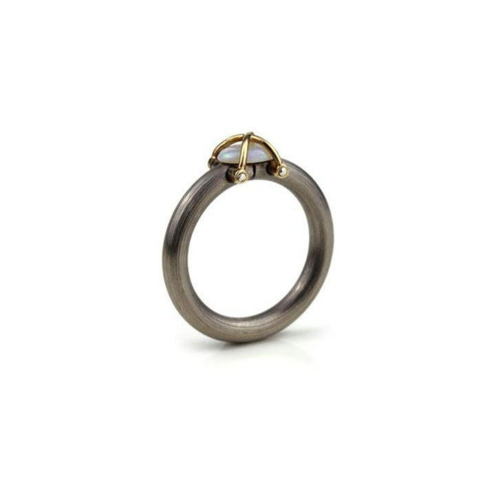 Une Opale en cabochon est présentée sur un anneau en acier inoxydable mat. Deux éléments d'or jaune entrecroisés et rivetés viennent entrelacer la pierre, comme une cage protectrice. Bijou de face.
