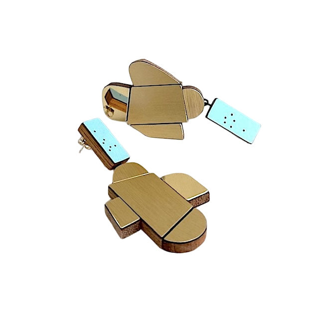 Suspendues à un support rectangulaire bleu clair à petits points noirs, ces boucles XL associent bois, formica doré texturé et doré poli façon miroir. Légères et sculpturales, elles sont faites à la main par l’artiste belge Karen Vanmol.