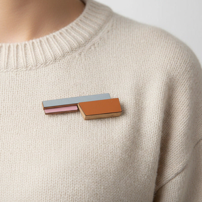 Broche géométrique allongée composée de trois éléments en bois recouvert de formica rose, gris clair et brun orangé. Bijou porté.