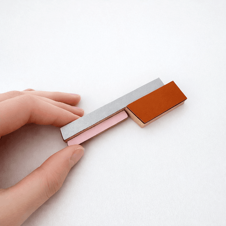 Broche contemporaine allongée en bois et formica gris,rose et brun orangé.  Détail du bijou tenu entre deux doigts. Pièce unique réalisée à la main par l’artiste belge Karen.