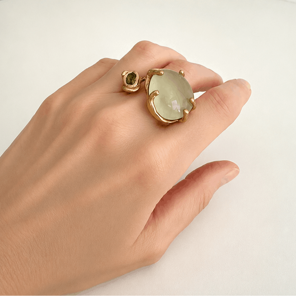 Main présentant une bague asymétrique, ouverte et sculpturale en bronze sertie d'une généreuse prehnite ronde et d'un péridot plus petit ovale et facetté.